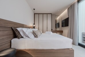 A modern hotel room with a wooden headboard, a large bed with white bedding, a bedside table, a lamp, a flat-screen TV mounted on the wall, and a view of the city through large windows. - Nysa Luxury Villa 5 ,Private Pool near Hersonissos (Heraklion)