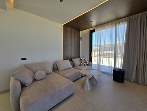 A modern living room with a sectional sofa, a round coffee table, and a view through large windows. - Nysa Luxury villa 5, Private pool, Near Hersonissos (Heraklion)