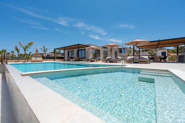 A swimming pool with lounge chairs and umbrellas, surrounded by modern architecture. - Nysa Luxury Villa 5 ,Private Pool near Hersonissos (Heraklion)