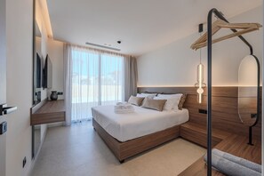 A modern hotel room with a large bed, a flat-screen TV, a wooden bedside table, a built-in bench, and a large window with curtains. - Nysa Luxury villa 6, Private pool, Near Hersonissos (Heraklion)