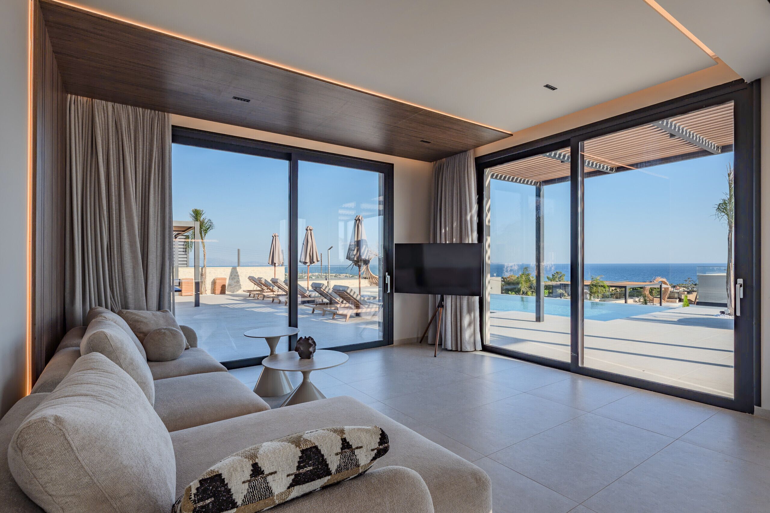A modern living room with a sofa, a flat-screen TV, and large sliding glass doors leading to a pool area with a view of the ocean.