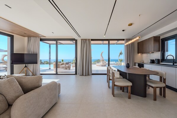 A modern living room with a sofa, a TV, a dining area with a table and chairs, and a kitchen with a large window. - Nysa Luxury villa 6, Private pool, Near Hersonissos (Heraklion)