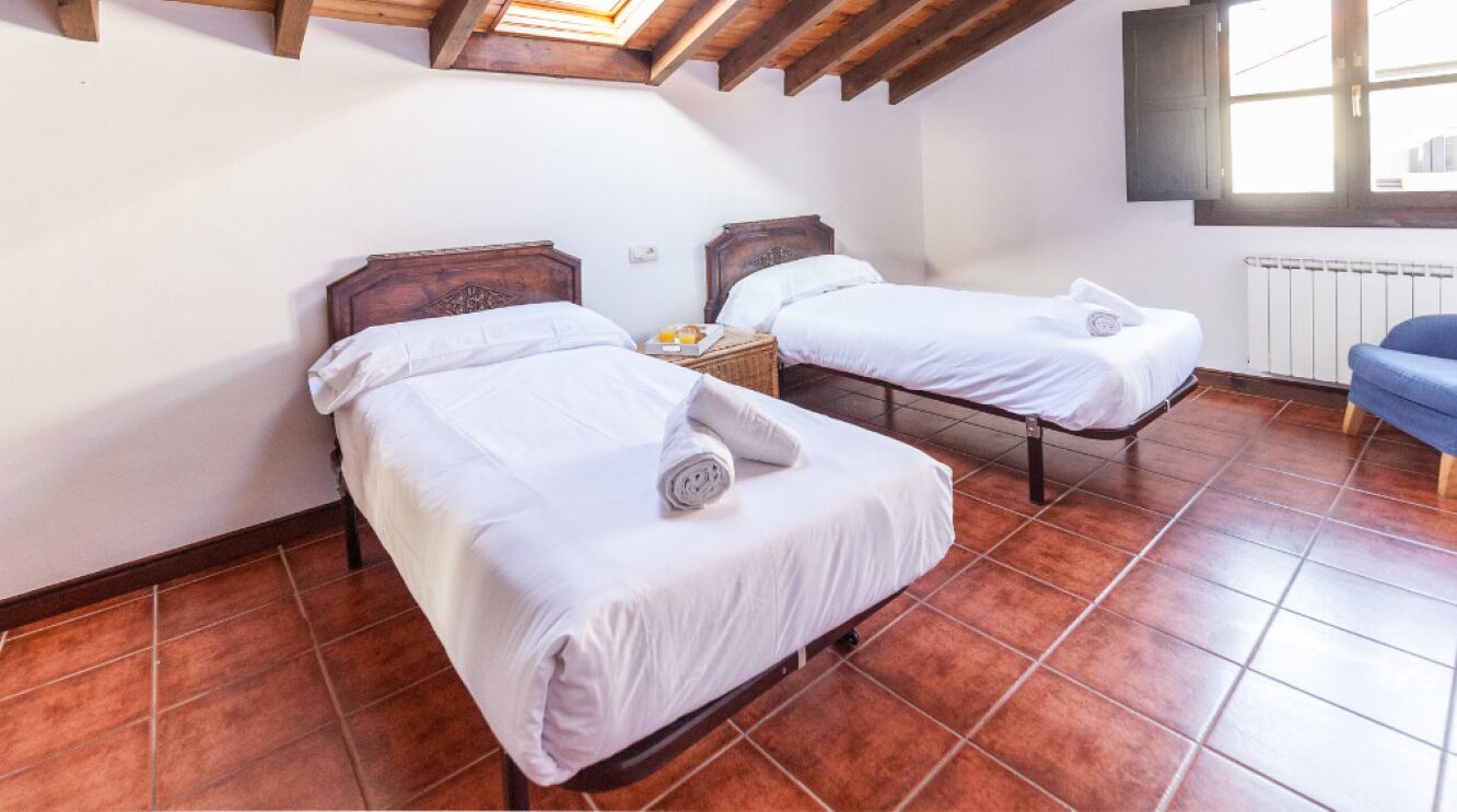 A room with two beds, a wooden ceiling, a radiator, and a window.
