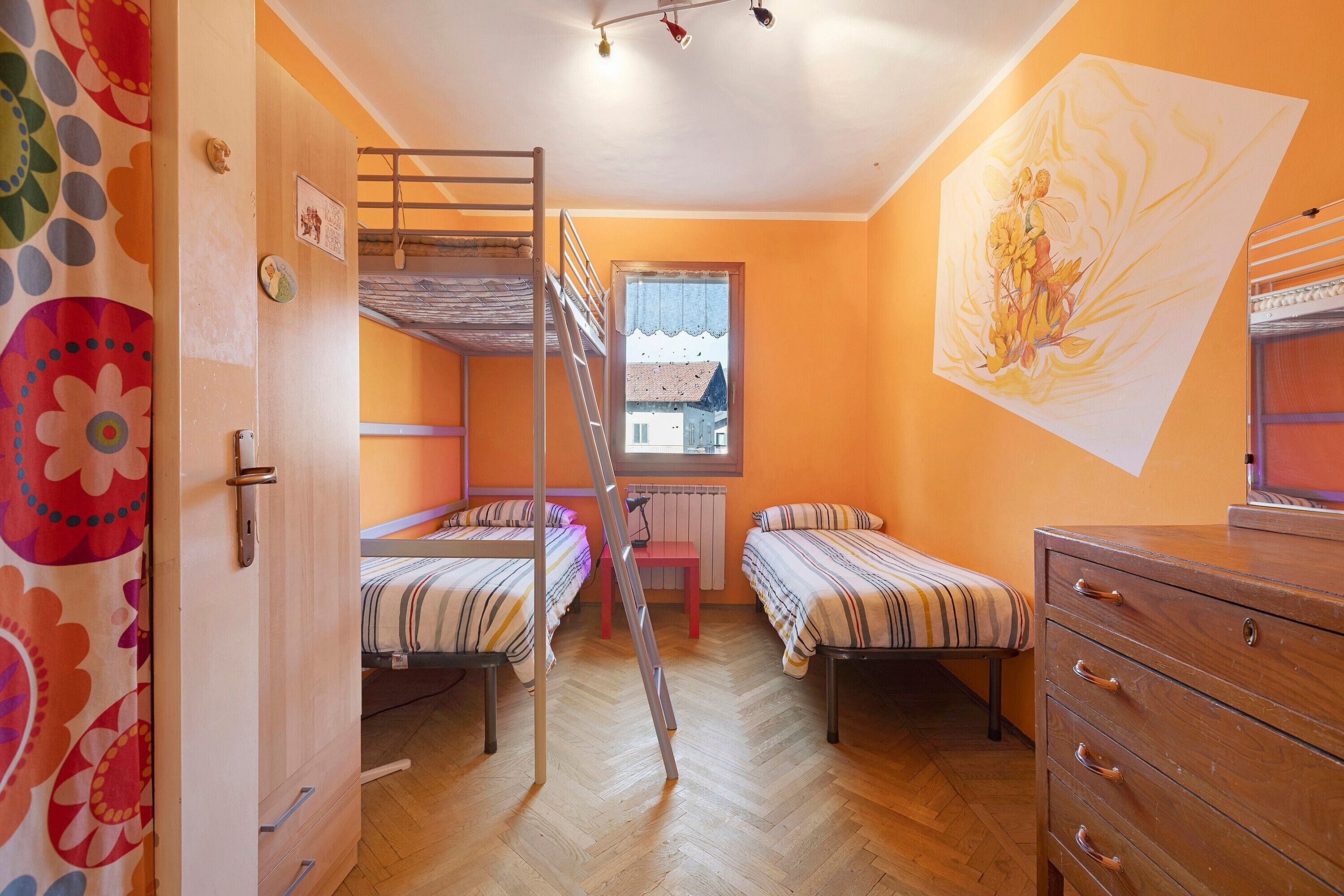 Une chambre de dortoir avec des lits superposés, une commode en bois, un radiateur et une fenêtre donnant sur des immeubles.