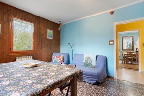 Ein Zimmer mit einer Holzwand, einer blauen Wand, einem Essbereich mit Tisch und Stühlen und einem Ausblick durch ein Fenster.