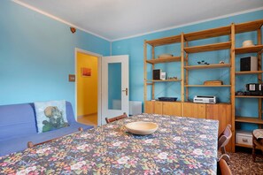 Ein Zimmer mit einer blumigen Tischdecke, einem blauen Sofa, einem Holzregal und einer Tür zu einem anderen Raum.