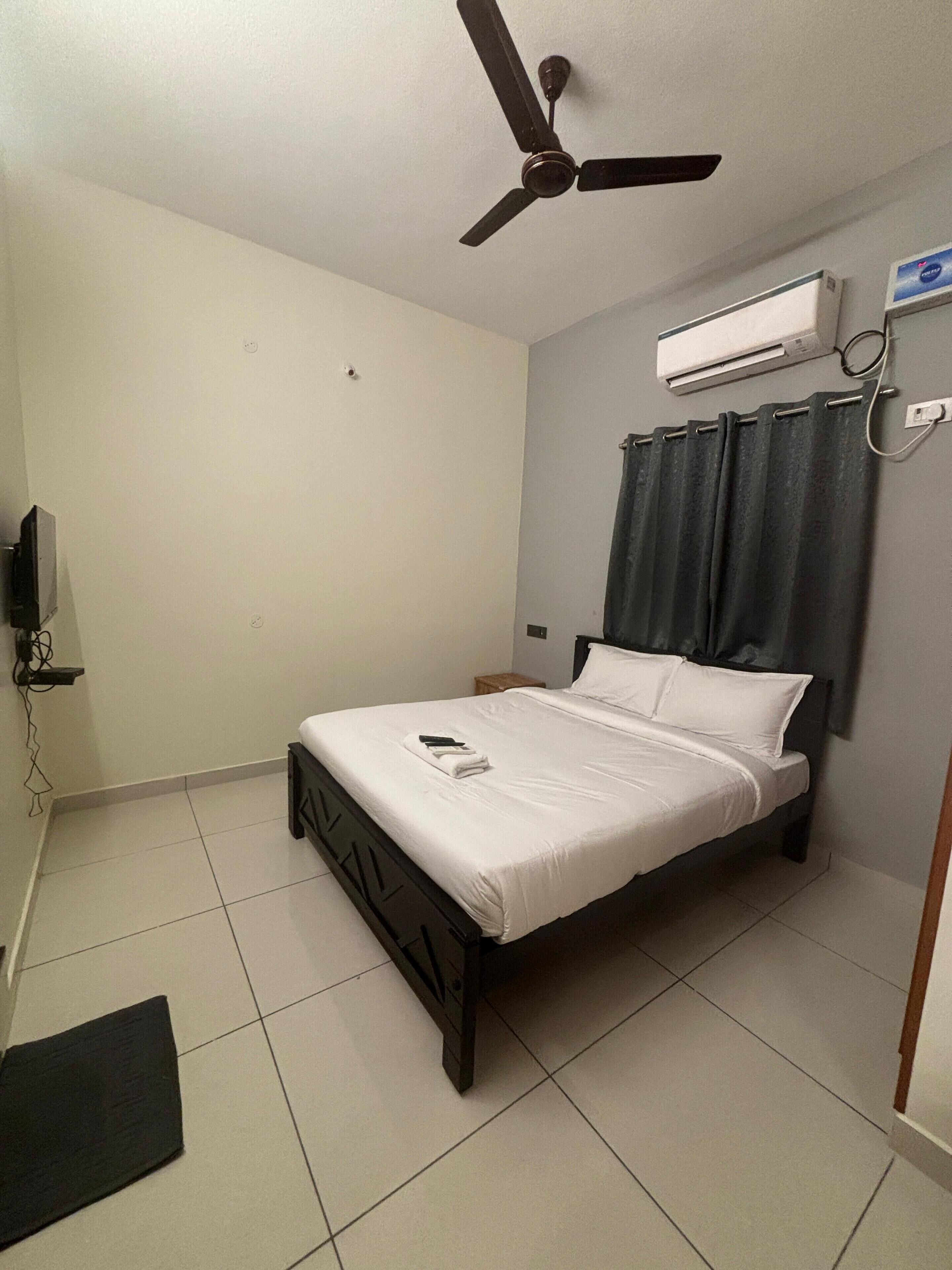 A bedroom with a bed, a ceiling fan, an air conditioning unit, and a TV mounted on the wall.