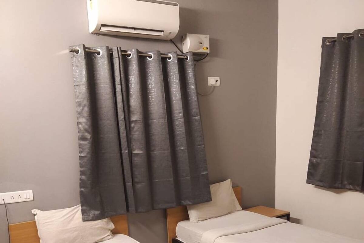 Camera d'albergo con due letti, condizionatore d'aria a parete, una tenda e un comodino con una bottiglia e telecomandi.