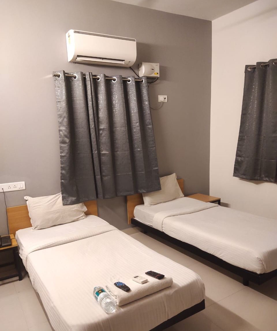 A hotel room with two single beds, an air conditioning unit, a curtain, and a nightstand with a bottle and remote controls.