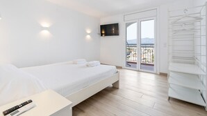 A modern bedroom with a bed, a TV mounted on the wall, a balcony with a railing, and a white shelving unit. - Welcs Apartamento 064 EMP Frente al Mar (Castello d'Empuries)