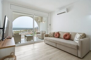 Ein modernes Wohnzimmer mit einer Couch, einem Fernseher und Blick auf den Strand durch eine Glastür.