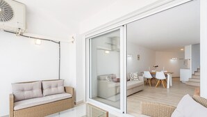 A modern living room with a glass coffee table, wicker sofas, and a sliding glass door leading to a dining area with a white table and chairs. - Welcs Apartamento 064 EMP Frente al Mar (Castello d'Empuries)