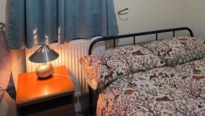 Un dormitori amb llit, una taula de nit amb una làmpada, un radiador i una cortina.
