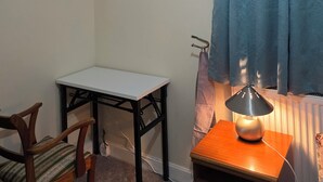 Una habitació petita amb sostre inclinat, una taula, una cadira, una làmpada i una finestra amb cortines blaves.