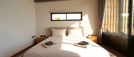 Quarto com cama grande, duas mesas de cabeceira, janela com persianas e vista para o verde lá fora.