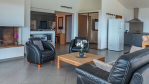 Una sala de estar moderna con sofás de cuero, una mesa de centro de madera y un televisor. Se ve una cocina con estufa y refrigerador al fondo.