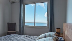 Un dormitorio con cama, una silla, una mesita de noche con lámpara y una ventana con vista al mar.