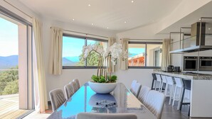 A modern kitchen with a large island, a dining area with a glass table, and a view of the mountains. - Villa Ghjiseppina Di Pinarello (Porto-Vecchio)