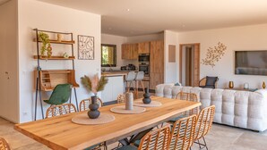 A modern kitchen with wooden cabinets, a dining area with a wooden table and wicker chairs, and a living area with a sofa and a TV. - Villa Felicita di Lecci (Lecci)