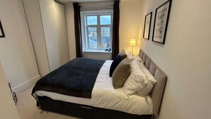 Une chambre d’hôtel moderne avec un grand lit, une fenêtre avec des rideaux, une lampe de chevet et des tableaux encadrés au mur.