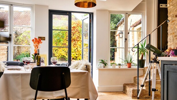 Un coin repas avec une table dressée pour deux personnes, un trépied et une vue sur un jardin, visible à travers de grandes fenêtres.