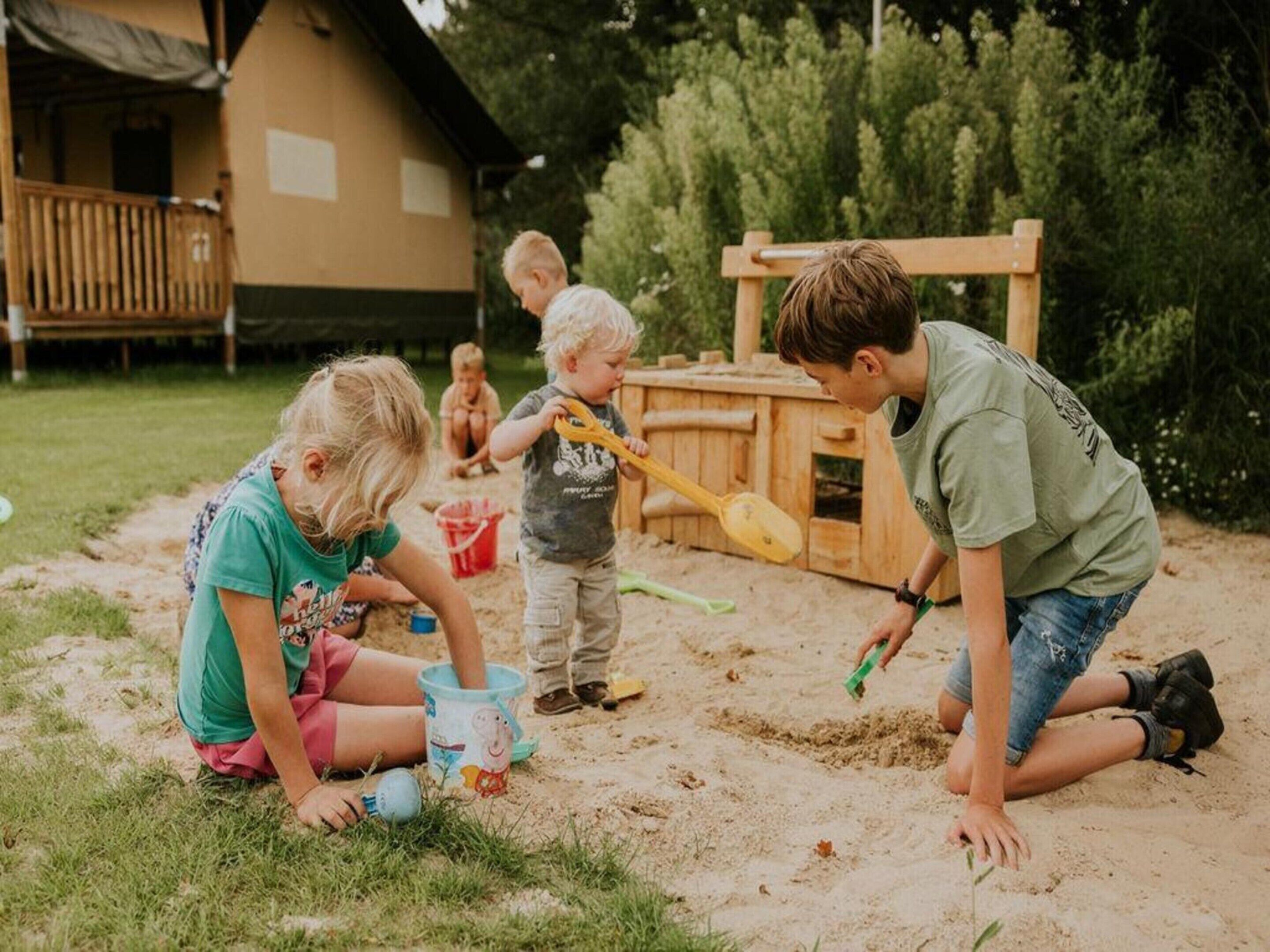 Children playing in a sandy outdoor area with a wooden playhouse in the background.