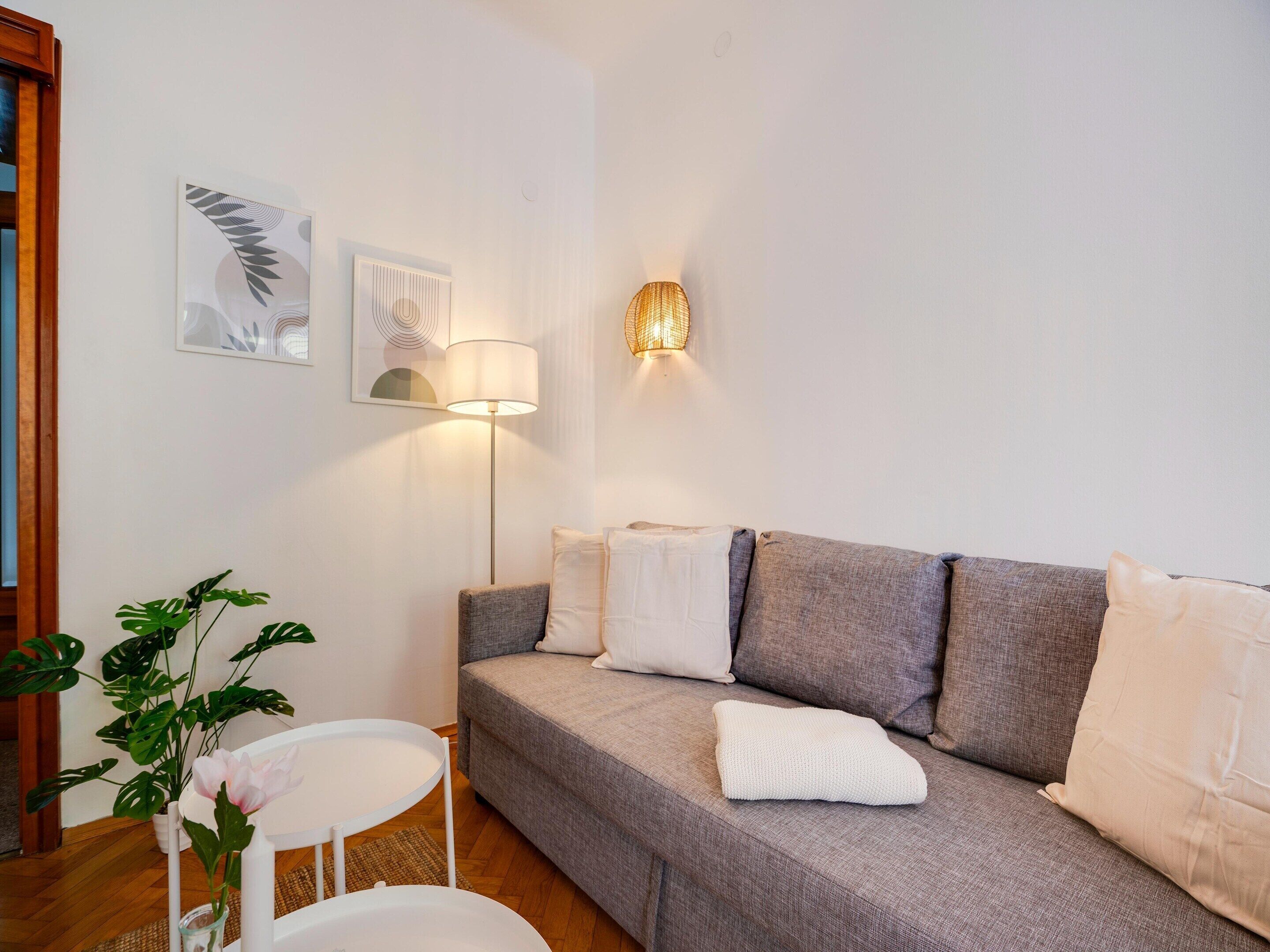 Ett modernt vardagsrum med en grå soffa, vita kuddar, en golvlampa och ett litet vitt bord med en krukväxt.