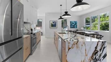 A modern kitchen with a marble island, stainless steel appliances, pendant lighting, and large windows.
