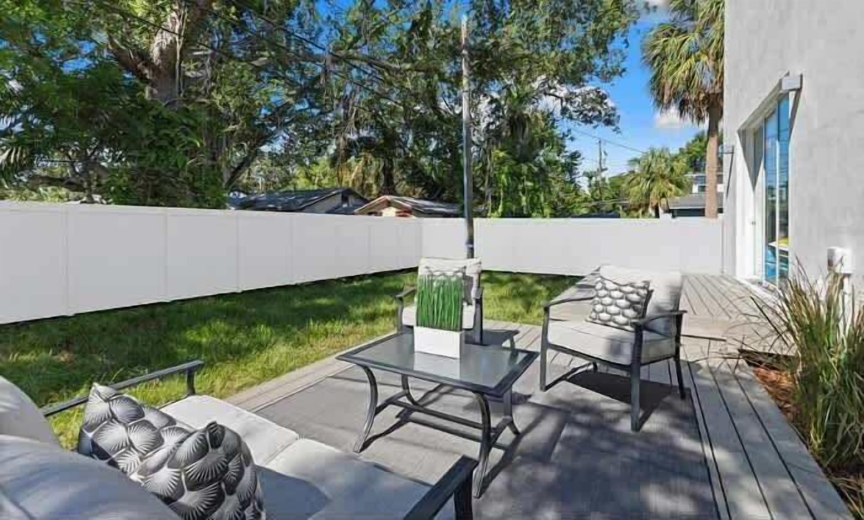 A modern outdoor patio area with a glass-top table and metal-framed chairs, surrounded by a white fence and lush greenery.