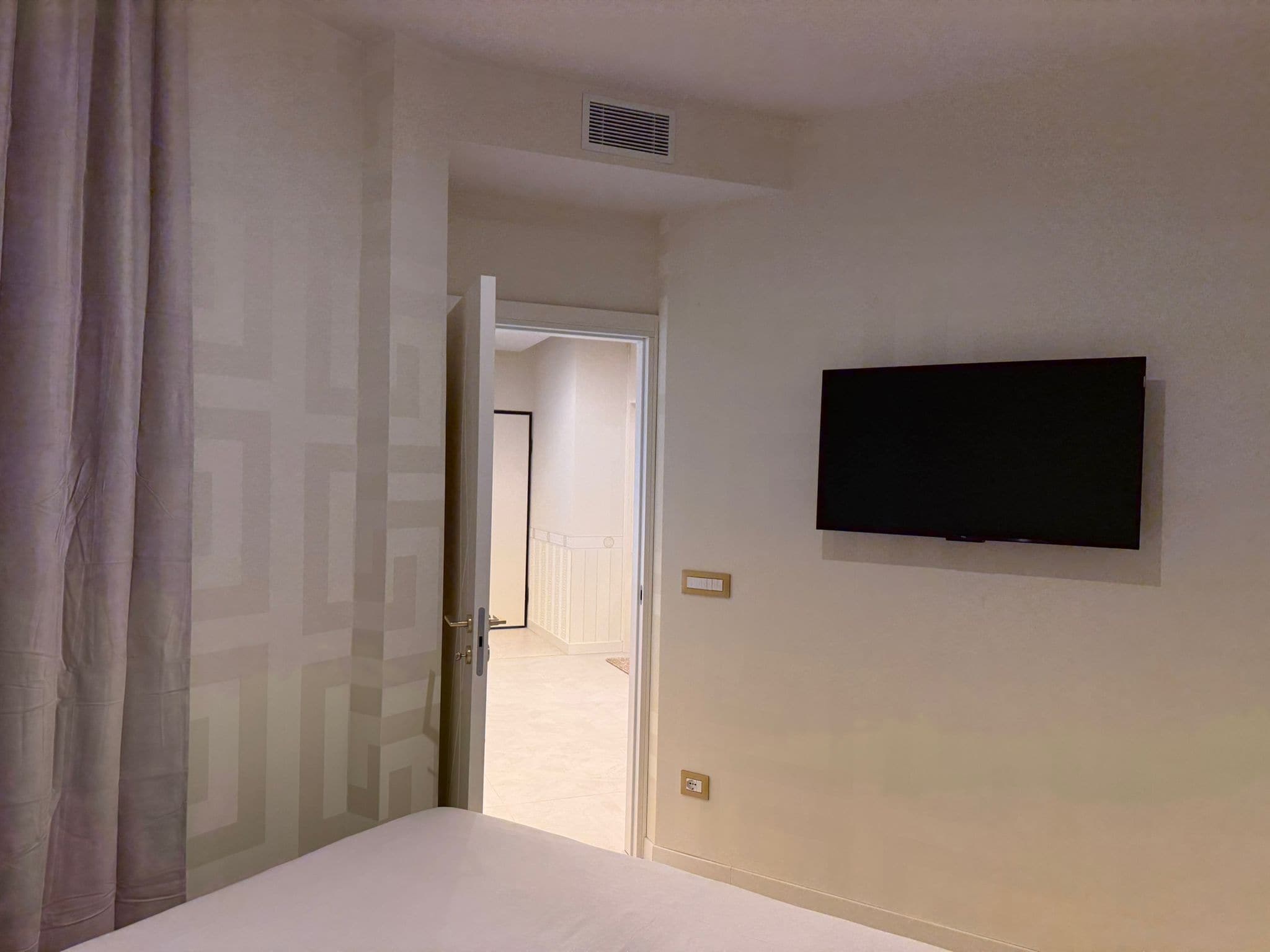 A hotel room with a bed, a television mounted on the wall, a door leading to another room, and a vent on the ceiling.