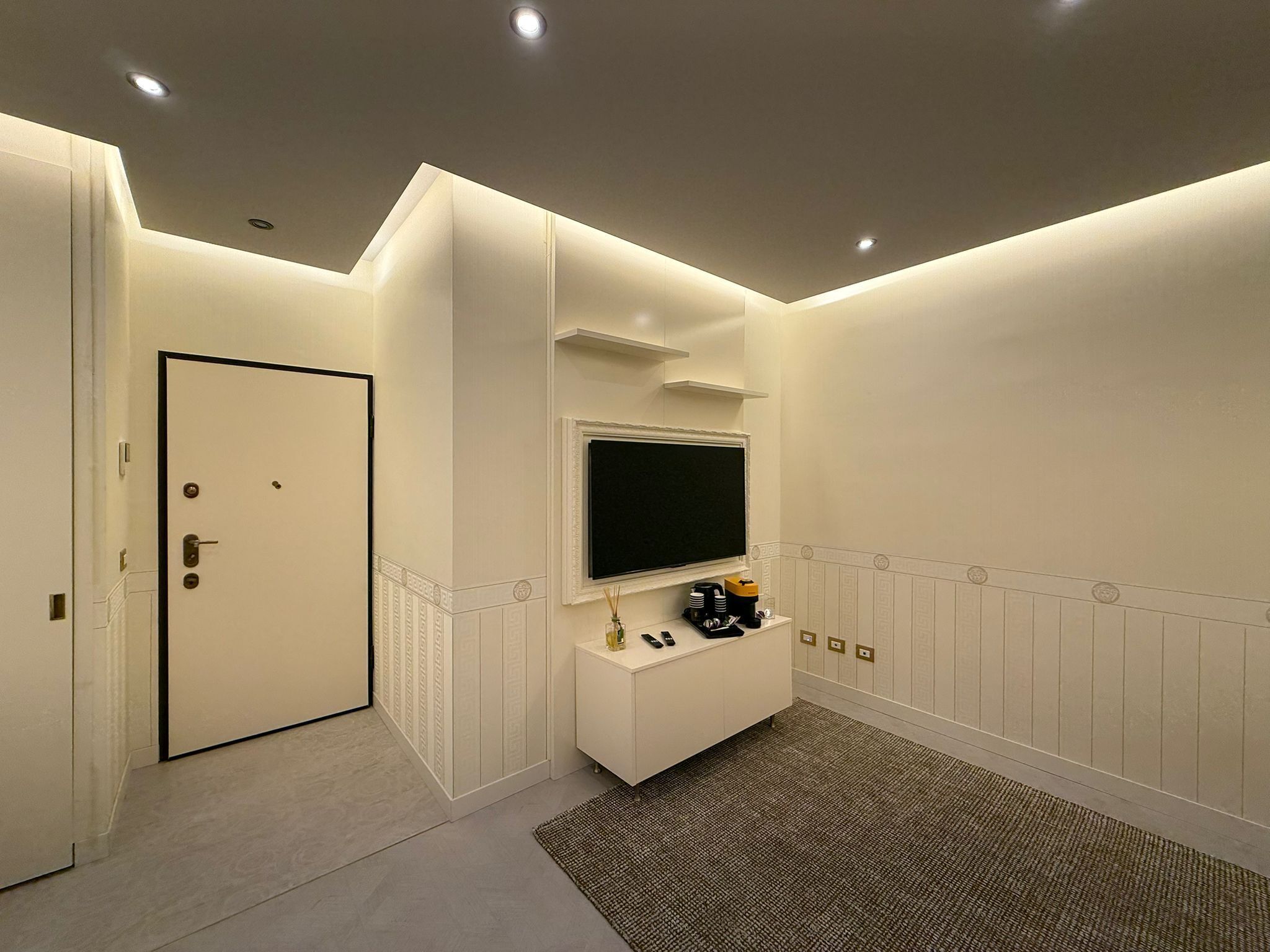 A modern, well-lit hallway with a door, a mounted TV, and a small cabinet with toiletries.