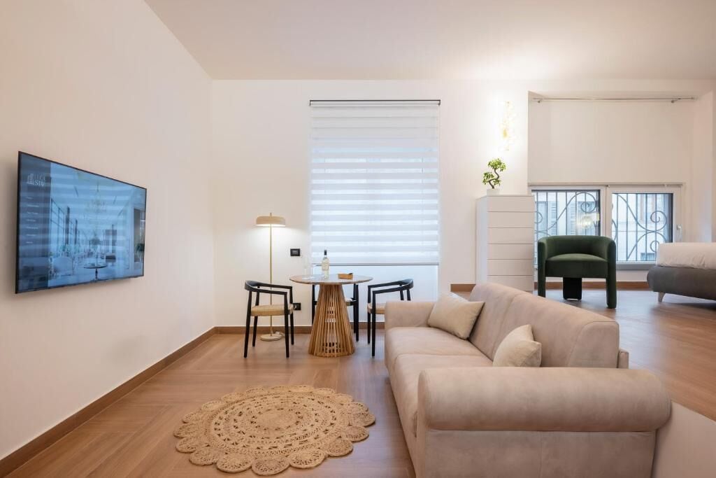 A modern living room with a beige sofa, a round coffee table, two chairs, a large framed picture, and a window with blinds.
