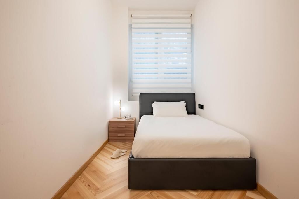 Une petite chambre à coucher moderne, avec un lit simple, une table de chevet, une lampe de chevet et une fenêtre avec des stores.