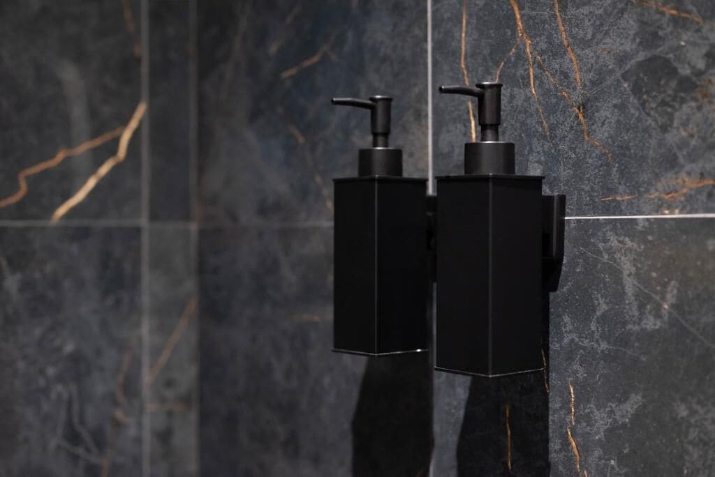 Deux distributeurs de savon noir sont fixés sur un mur recouvert de carreaux de marbre foncé.