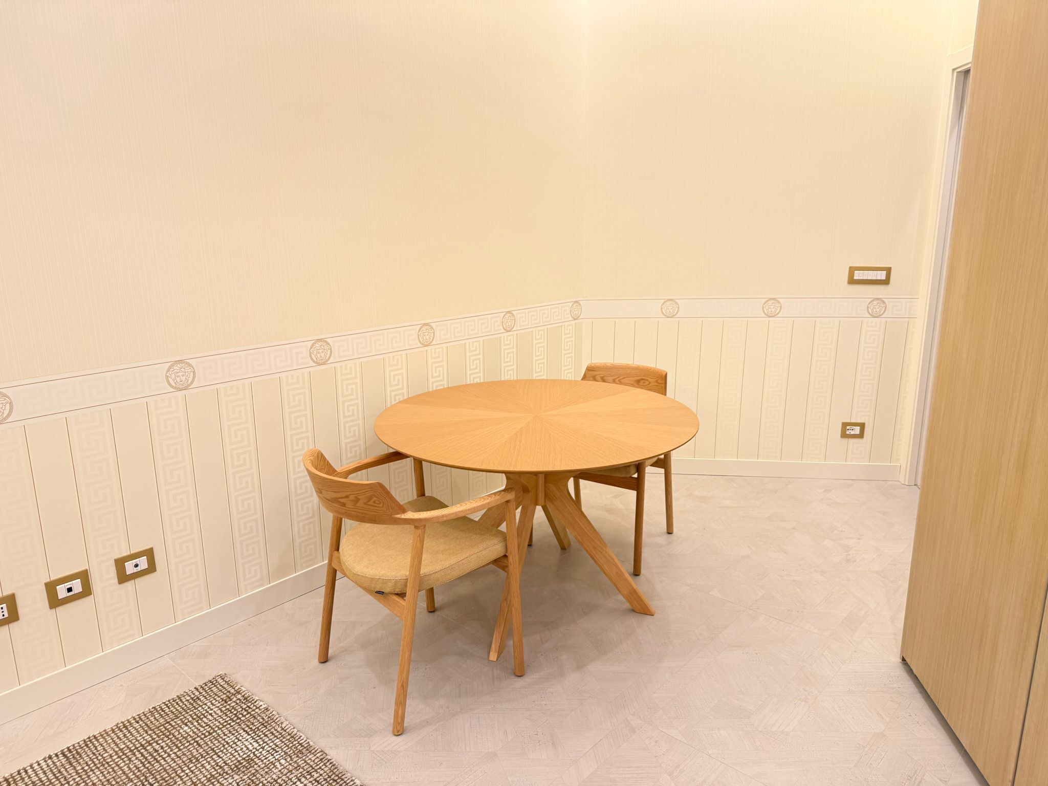 Uma pequena área de jantar com uma mesa de madeira redonda e quatro cadeiras, parede com padrão decorativo e uma porta.