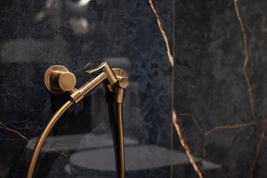 Chuveiro com design curvo, fixado em parede de mármore escuro com veios aparentes.