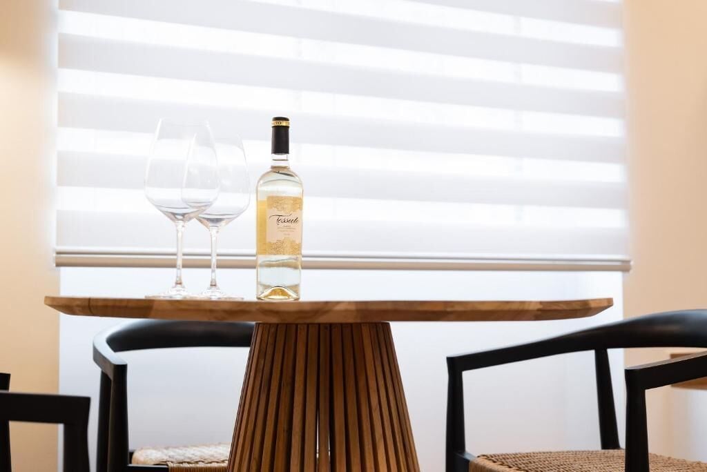 A table with a bottle of wine and two glasses, set against a window with blinds.