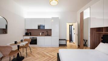 A compact hotel room with a kitchenette, a dining area with a round table and chairs, a bed, and a large mirror.