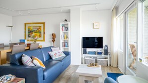 A modern living room with a blue sofa, a flat-screen TV, a bookshelf, and a dining area with a table and chairs. - 2 Person Apartment With sea View (De Koog)