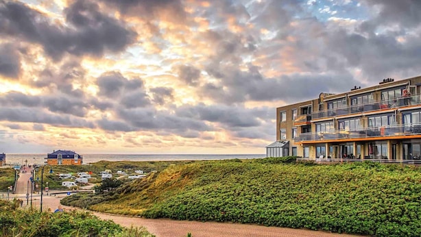 A coastal hotel with multiple balconies overlooking the sea, a paved walkway, and a landscaped area with dense greenery. - 2 Person Apartment With sea View (De Koog)