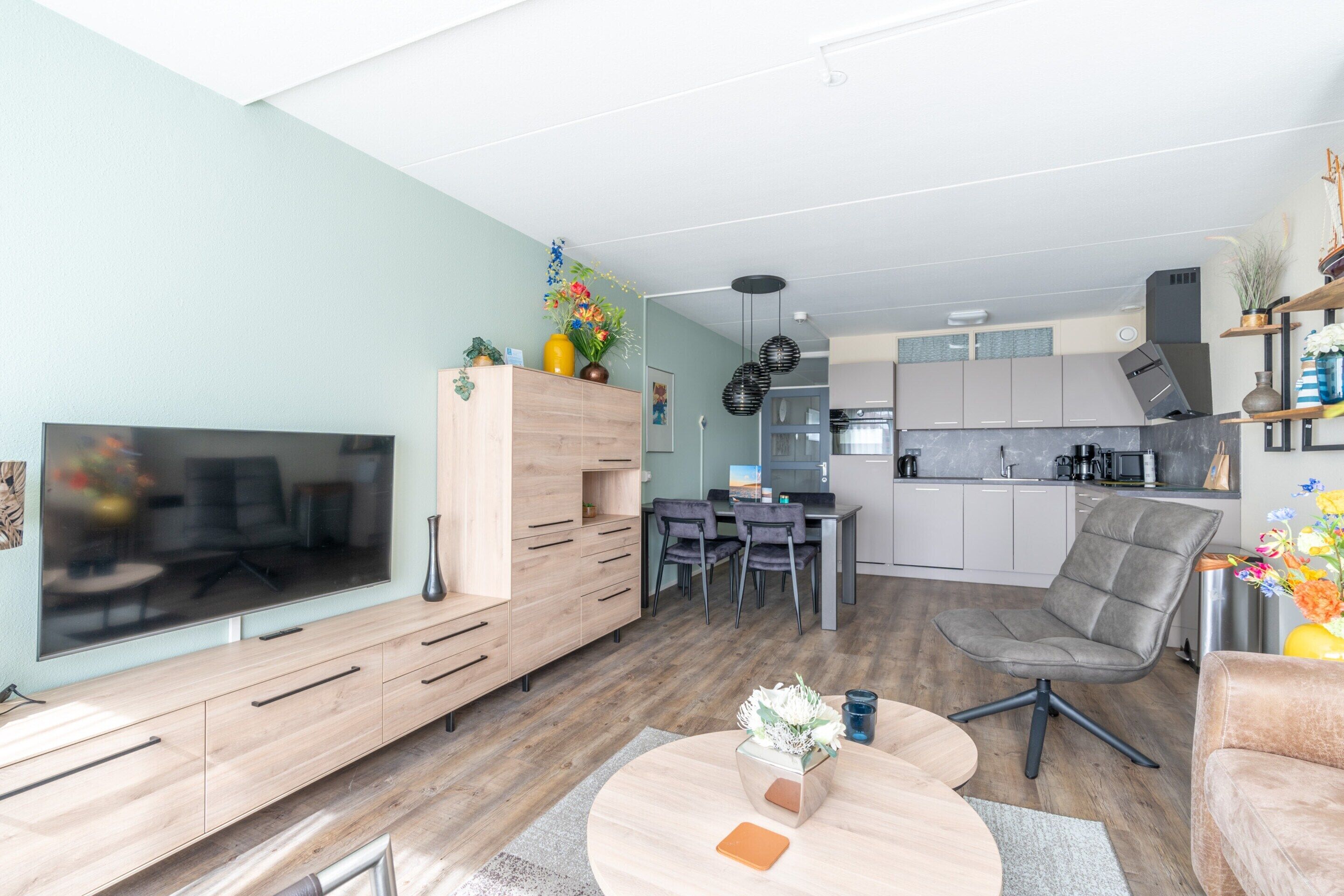 A modern living room with a flat-screen TV, wooden entertainment unit, and a dining area with a table and chairs.