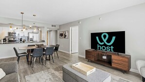 A modern living room with a dining area, a kitchen, and a TV displaying the word "host". - 1BR Retreat | Free Parking | Rooftop Pool (Miami)