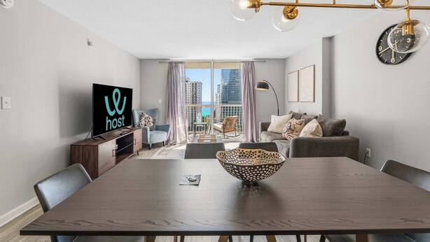 A modern living room with a dining table, a TV on a wooden cabinet, a large window with curtains, and a city view. - 1BR Retreat | Free Parking | Rooftop Pool (Miami)