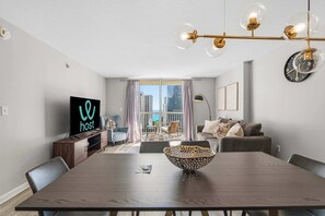 Ein modernes Wohnzimmer mit Esstisch, einem Fernseher auf einer Holzkommode, einem großen Fenster mit Vorhängen und Blick auf die Stadt.