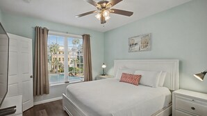 Un dormitorio con cama, mesitas de noche, ventilador de techo, ventana con cortinas y vistas a edificios.