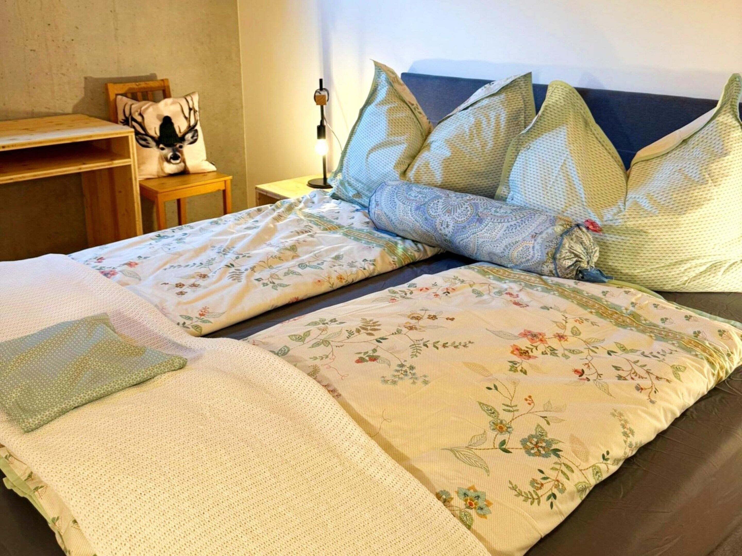 Un lit bien fait, avec une literie à motifs floraux et plusieurs oreillers, une table de chevet avec une lampe, et une chaise avec un coussin.