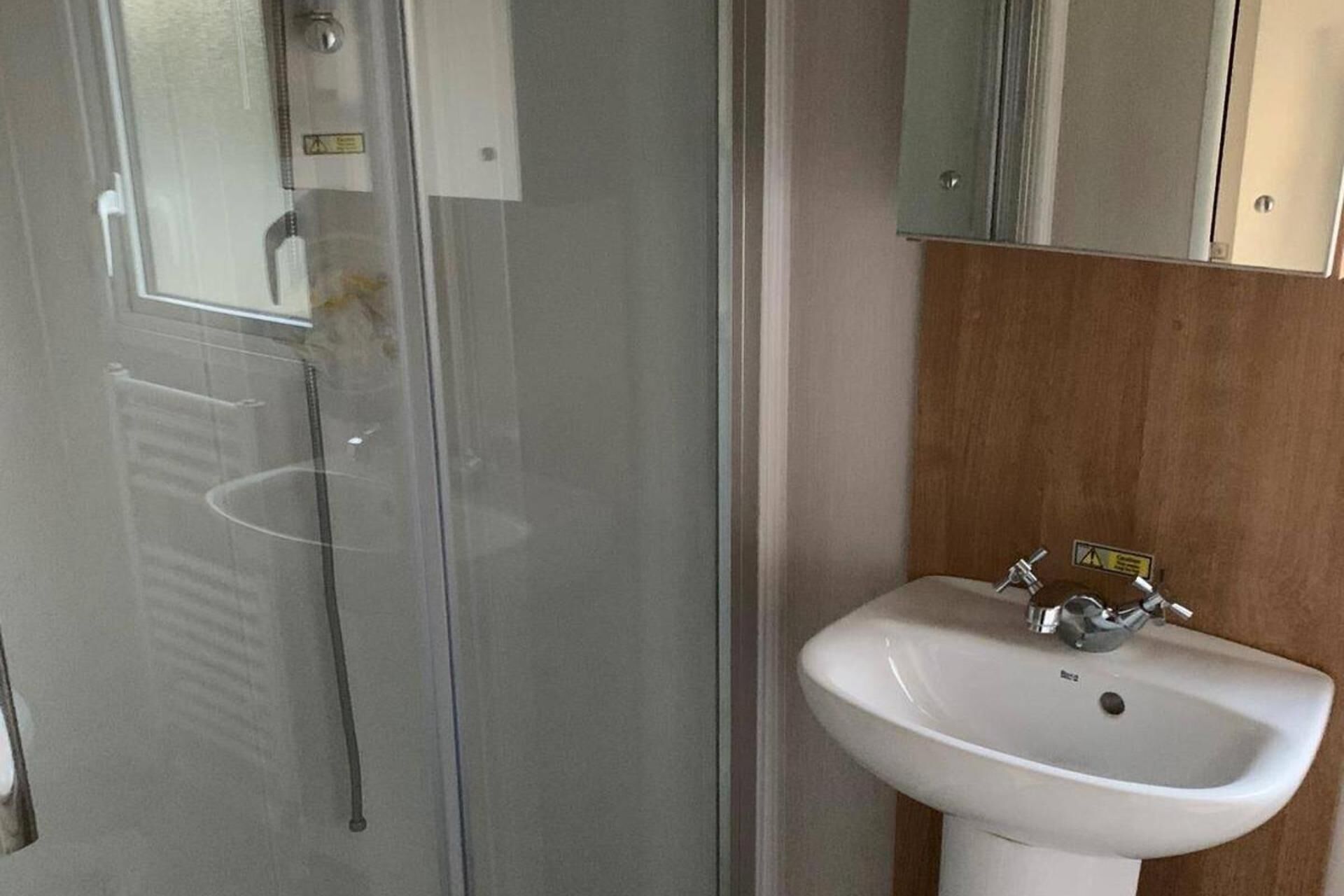 A bathroom with a glass shower enclosure, a white sink with two faucets, and a wooden panel wall.