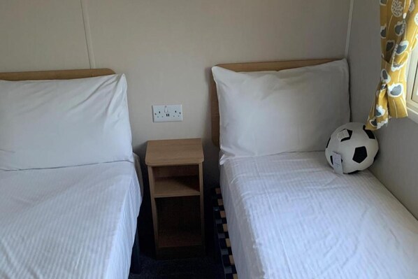 Deux lits simples avec du linge de lit blanc, une petite table de chevet en bois et un ballon de foot sur le lit de droite.