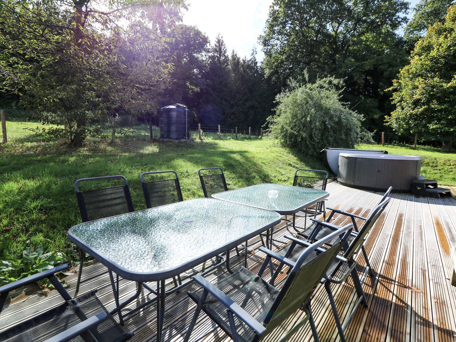 A wooden deck with a glass-top table and metal chairs, surrounded by a grassy yard and trees.