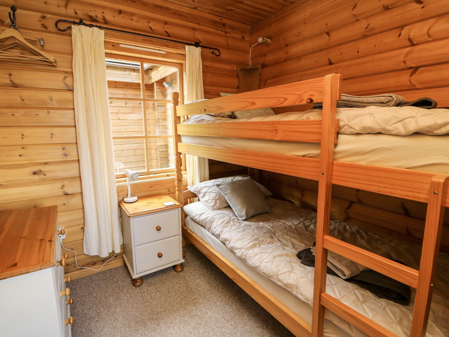 A wooden cabin room with bunk beds, a bedside table with a lamp, and a window with curtains.